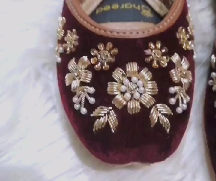 A product showcase video of maroon velvet jutti shoes on a rug surface. The video captures close-up details of the handcrafted gold floral embroidery and pearl accents from multiple angles, highlighting the shape, premium velvet texture, and cushioned inner soles.