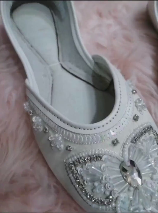 White bridal flats with crystal, sequin and pearl embellished butterfly design shown in close-up on a soft pink background.