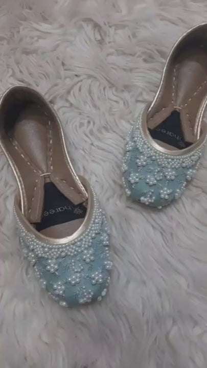 Close-up video of sky blue bridal juttis with floral imitation pearls, silver embroidery and tan insoles on a pale textured rug.